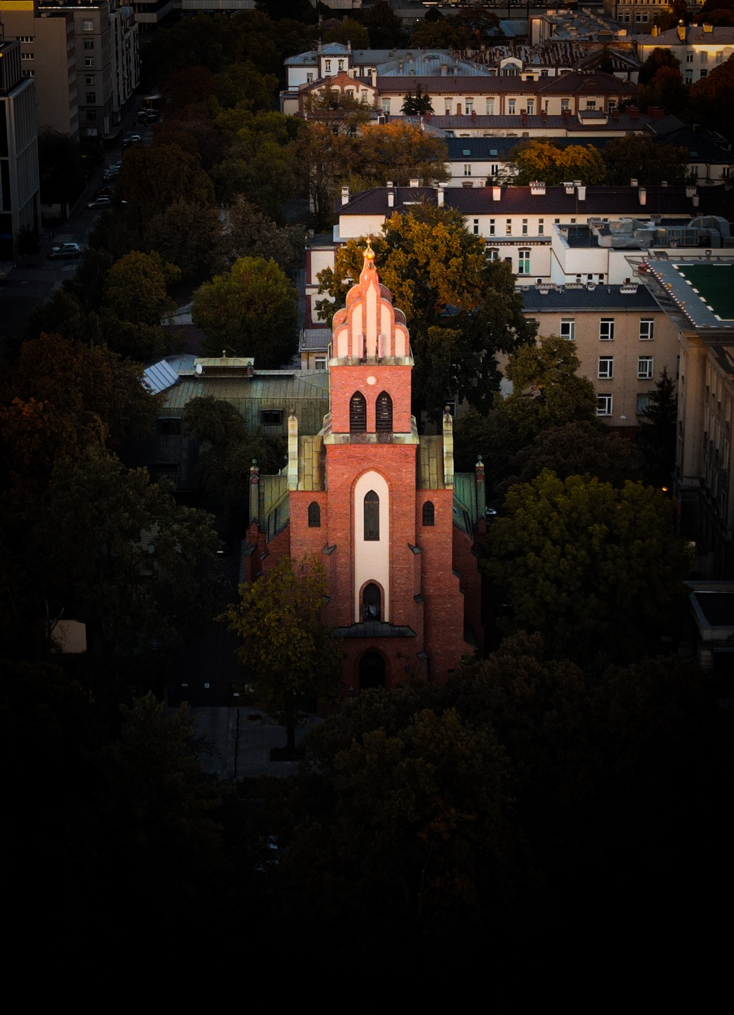 Widok z góry na ceglany kościół z wieżą, otoczony drzewami, na tle miejskiego krajobrazu z budynkami w jesiennej scenerii.