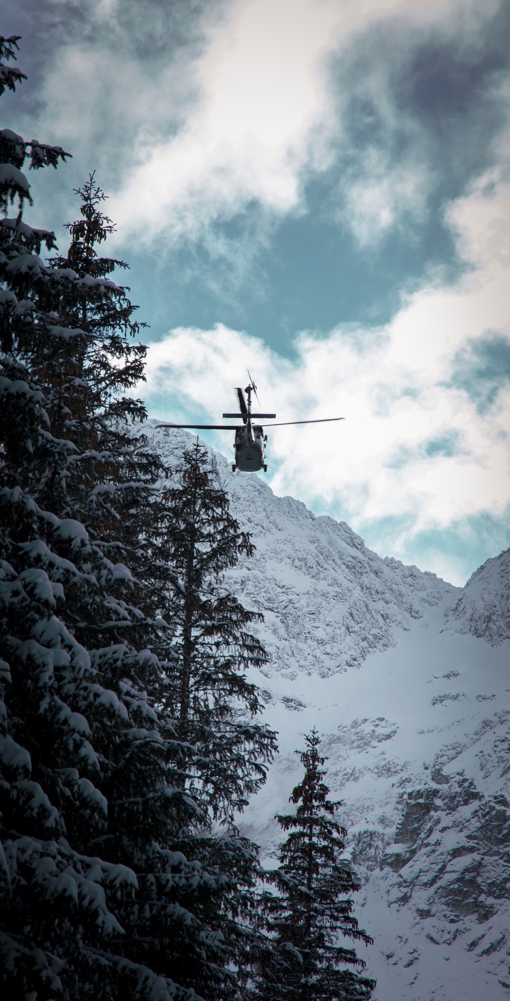 Helikopter w locie na tle ośnieżonych gór i lasu iglastego, błękitne niebo z chmurami w tle.