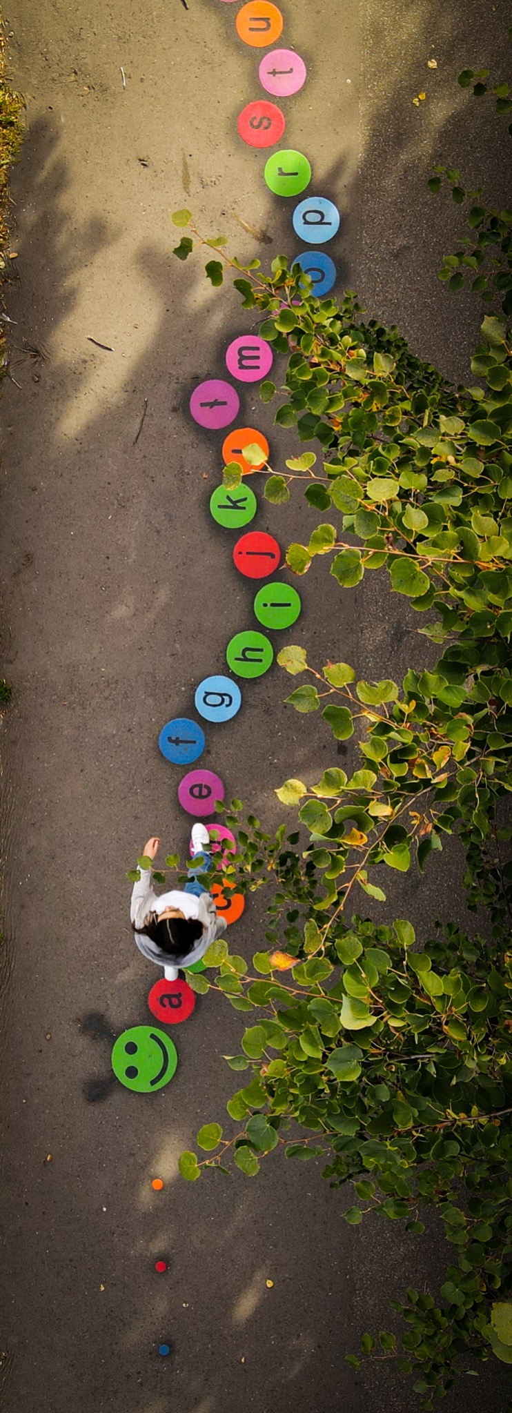 Dziewczynka widziana z góry, stojąca na kolorowych okrągłych płytach z literami alfabetu, tworzących ścieżkę edukacyjną na szarym podłożu, obok zielonych gałęzi drzew.