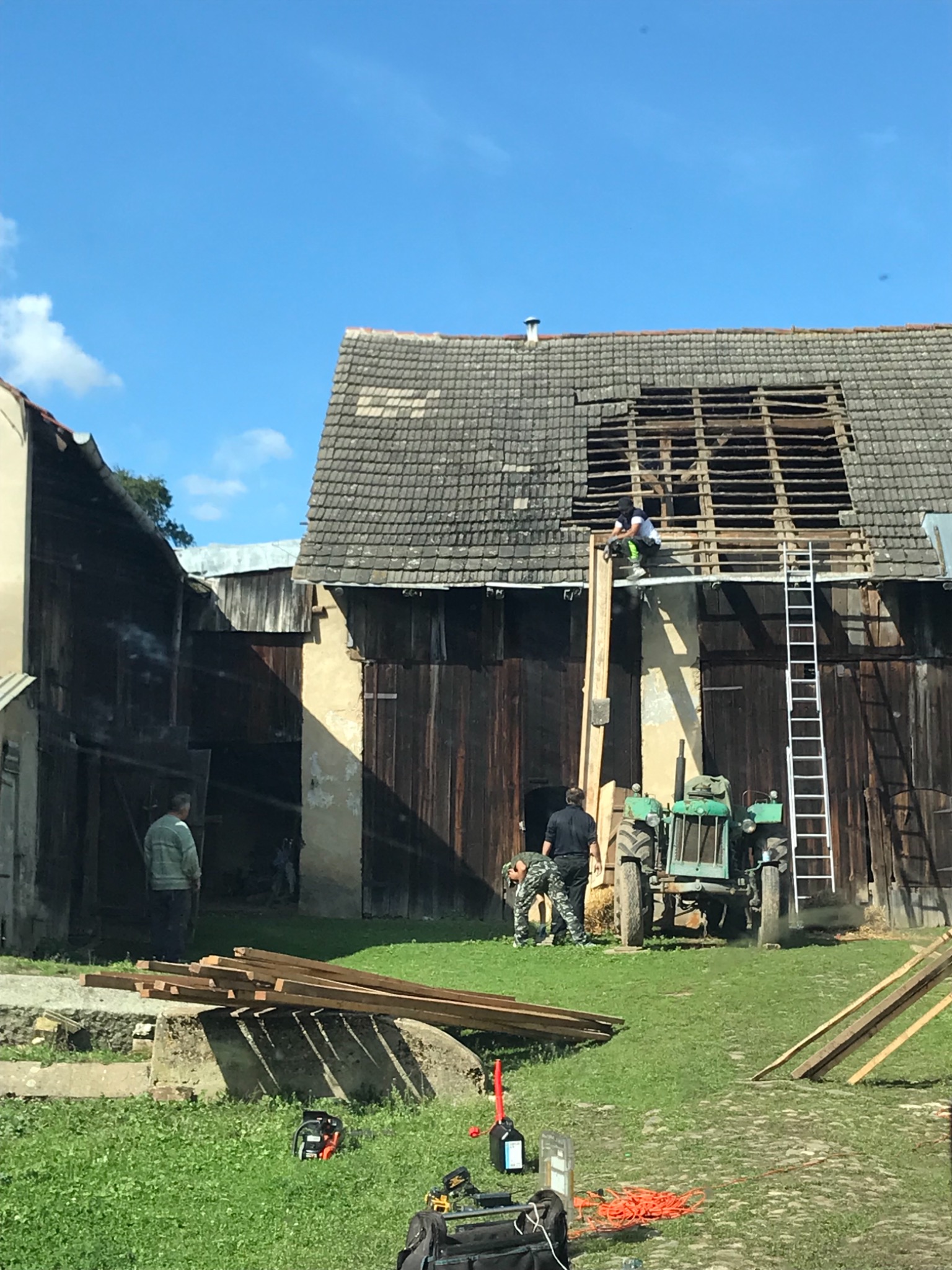 Demontaż starego dachu z eternitu na budynku gospodarczym, widoczny pracownik na dachu, drabina, traktor i narzędzia na trawie.