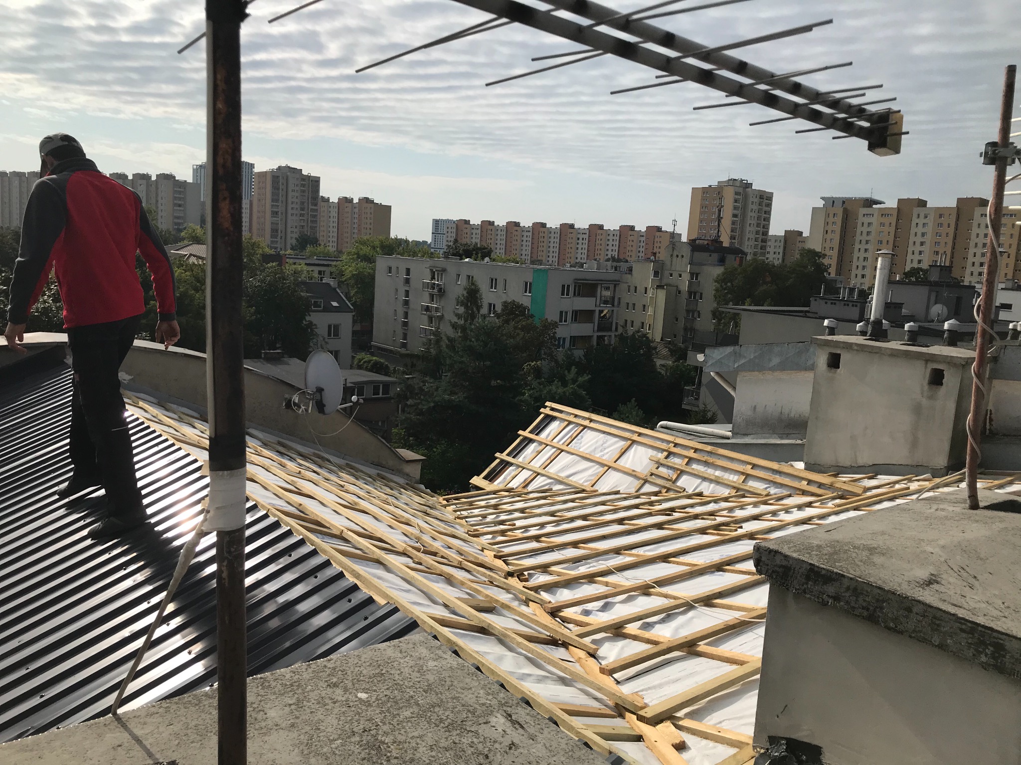 Pracownik w czerwonej bluzie chodzi po metalowym dachu w trakcie budowy, widoczne drewniane łaty i membrana, w tle zabudowa miejska i pochmurne niebo.