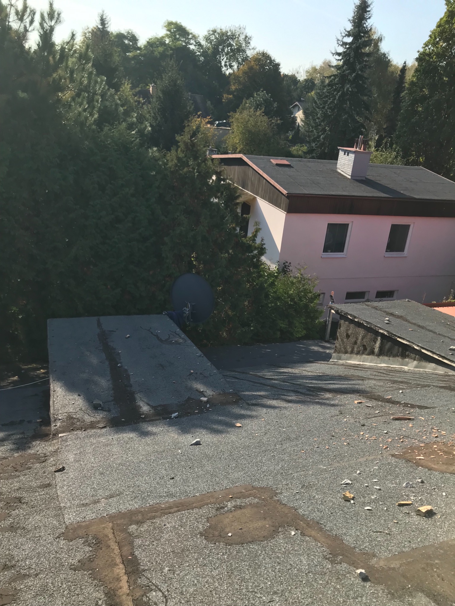 Uszkodzony, płaski dach pokryty papą, z widocznymi fragmentami gruzu i pozostałościami po pracach dekarskich, w tle dom z płaskim dachem i drzewami.