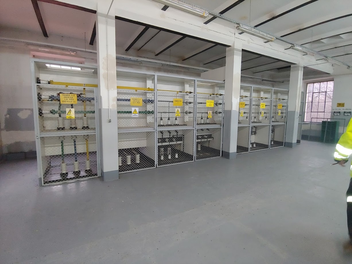 Wnętrze rozdzielni elektrycznej z zabezpieczonymi klatkami transformatorami 20/10 kV, widoczne tabliczki ostrzegawcze i instalacje elektryczne.