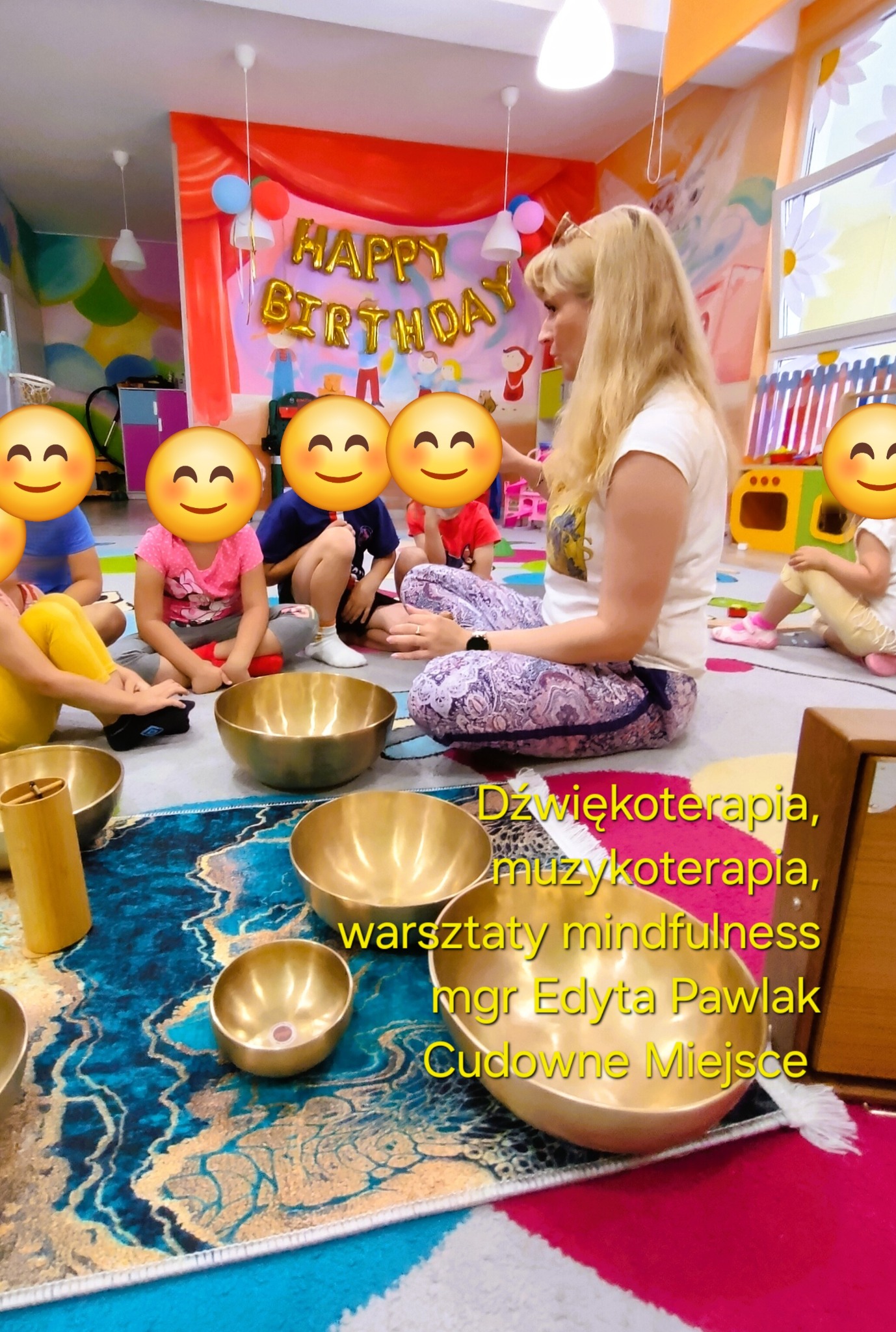 Zajęcia muzykoterapii dla dzieci z terapeutką. Dzieci siedzą na dywanie wokół mis dźwiękowych. W tle dekoracja urodzinowa i kolorowe ściany.
