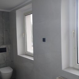 Glazura Damian Grendys - Wykończona łazienka w szarościach: płytki imitujące marmur na ścianach, grafitowa podłoga, biała toaleta podwieszana i okno z białymi ramami.