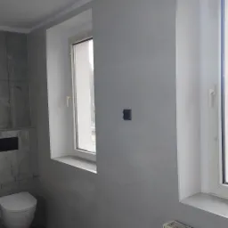 Wykończona łazienka w szarościach: płytki imitujące marmur na ścianach, grafitowa podłoga, biała toaleta podwieszana i okno z białymi ramami.