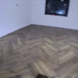 Ułożone panele podłogowe w jodełkę w narożnym pokoju z białymi ścianami i oknem zabezpieczonym folią.