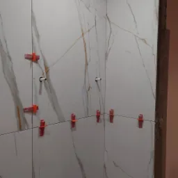 Montaż płytek imitujących marmur na ścianie z użyciem czerwonych klipsów poziomujących, widoczne rury instalacyjne, surowa ościeżnica drzwi