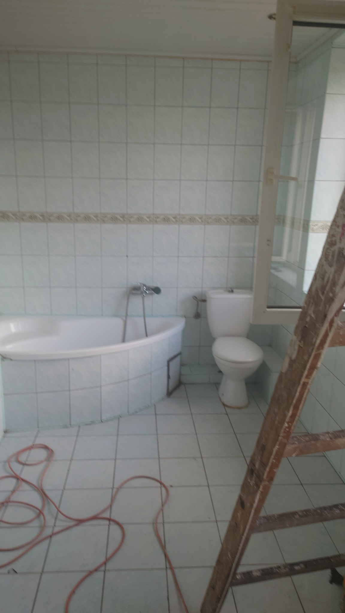 Jasna łazienka w trakcie remontu: biała wanna, toaleta i drabina malarska. Widoczne płytki na ścianach i podłodze. Kabel elektryczny leży na podłodze.