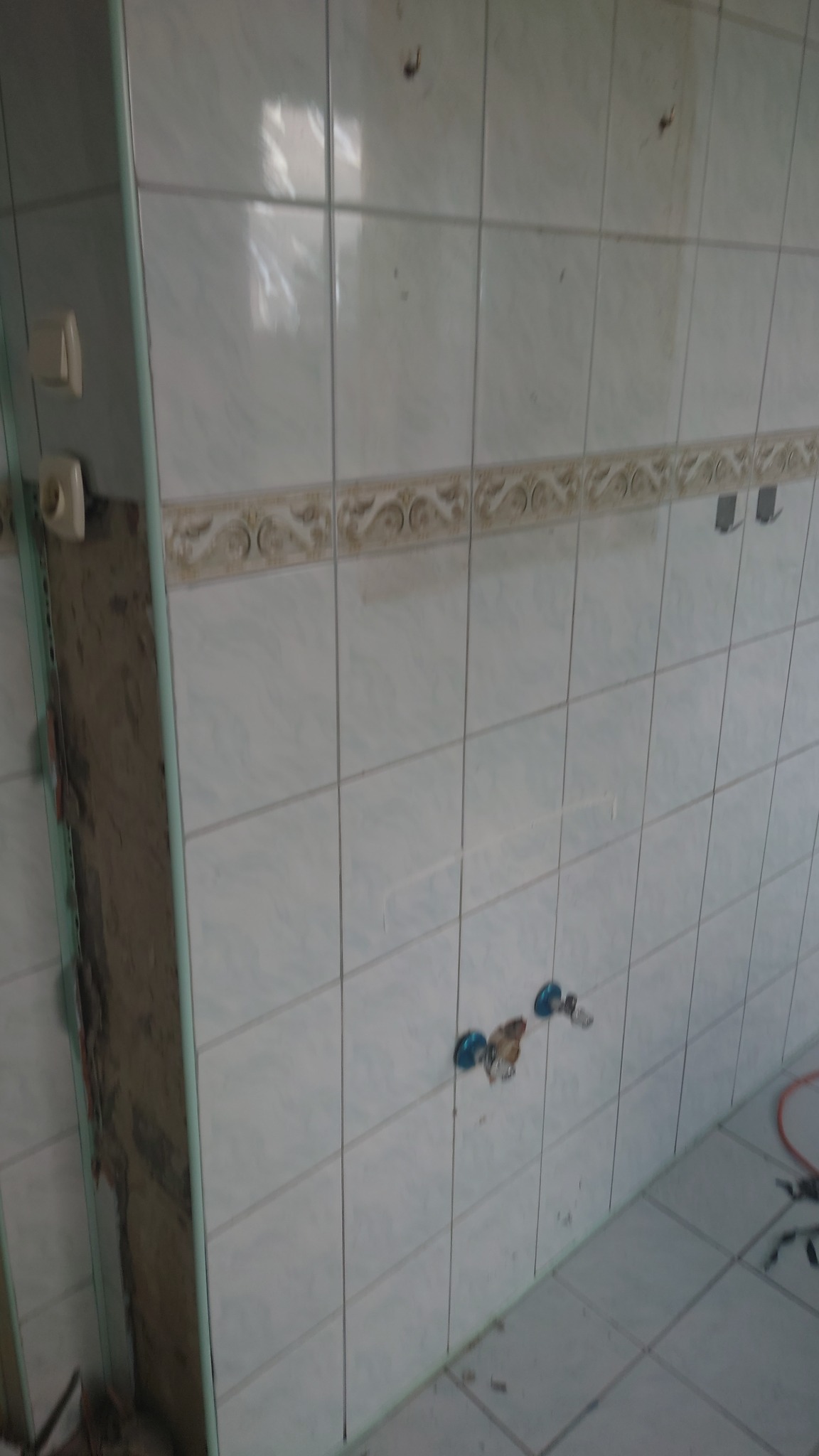 Ściana z częściowo usuniętymi, jasnymi płytkami ceramicznymi, widoczne przyłącza wodne i fragment instalacji elektrycznej. Fragment ściany bez płytek, w trakcie prac przygotowawczych.