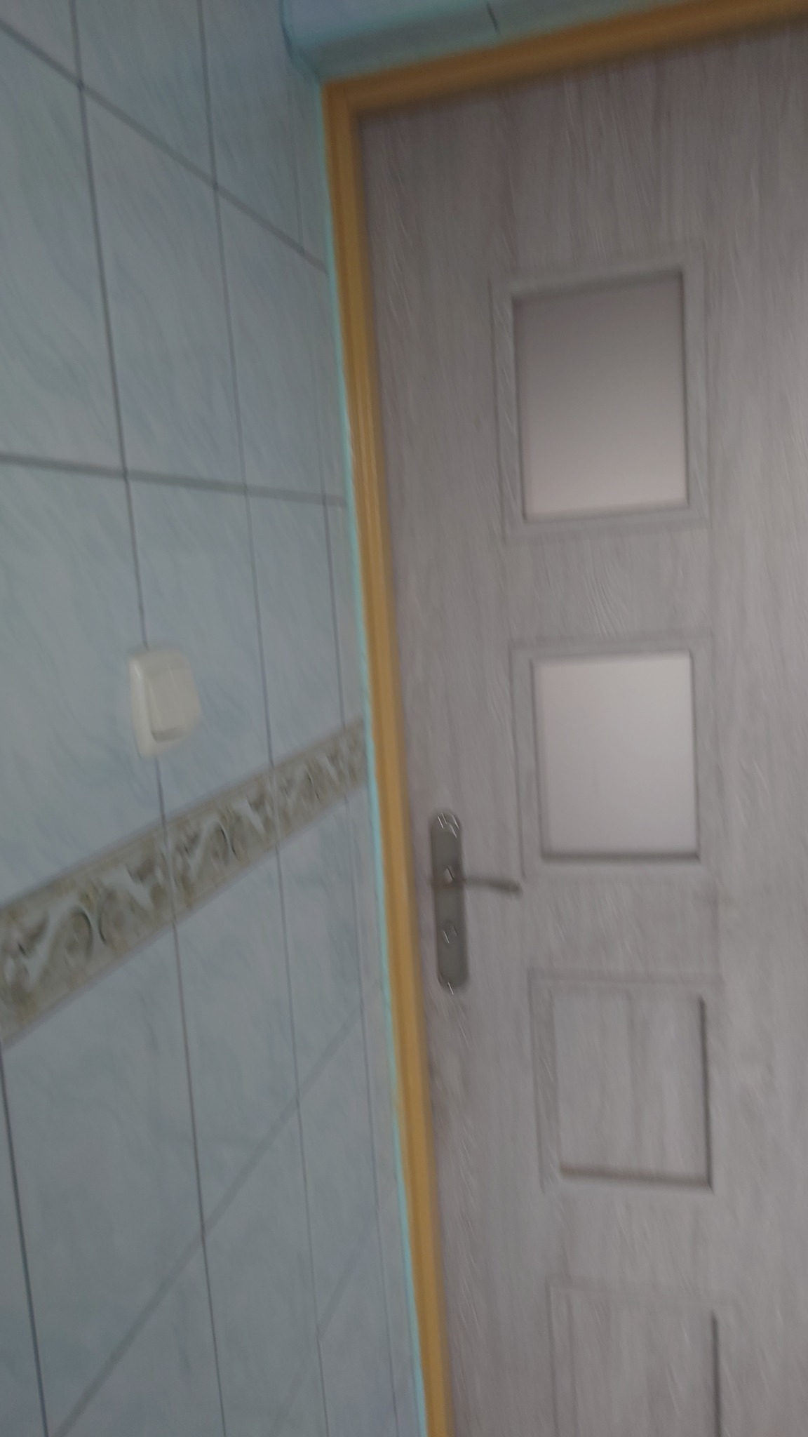 Błękitna ściana z dekoracyjną glazurą sąsiaduje z jasnymi drzwiami z trzema szybkami, wyposażonymi w srebrną klamkę. Widoczny włącznik światła.