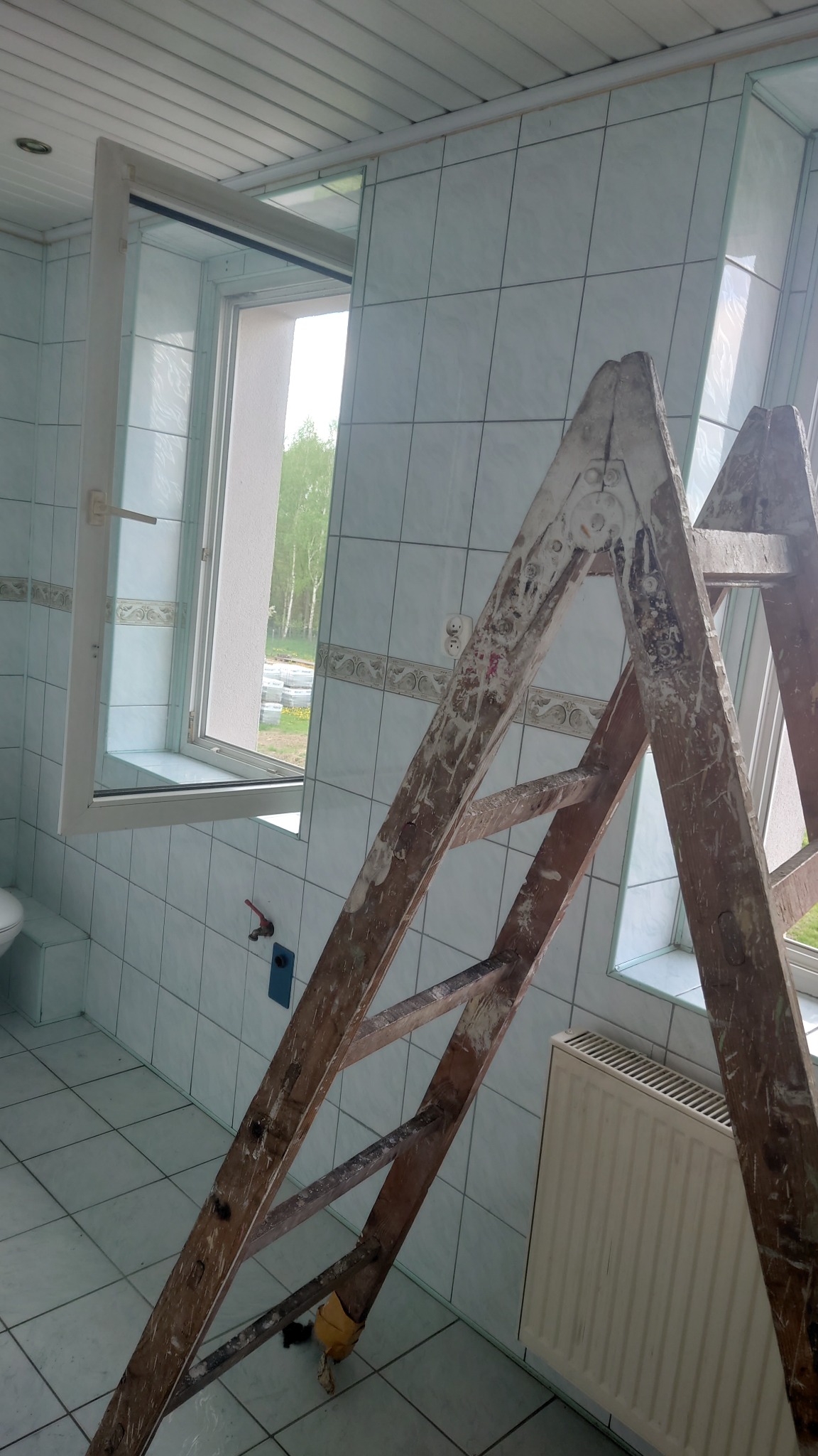 Łazienka w trakcie prac: białe płytki, okno z widokiem na zieleń, drewniana drabina z resztkami farby, grzejnik. Widoczny etap prac wykończeniowych.