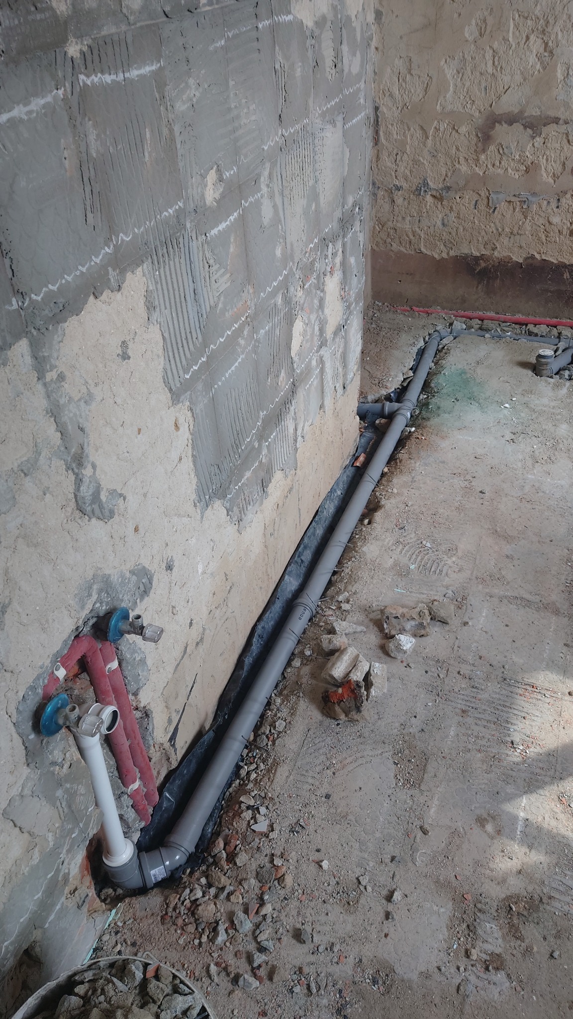 Szare rury kanalizacyjne ułożone wzdłuż ściany z resztkami zaprawy, czerwone rury wystają ze ściany. W tle gruz i pozostałości po pracach budowlanych.