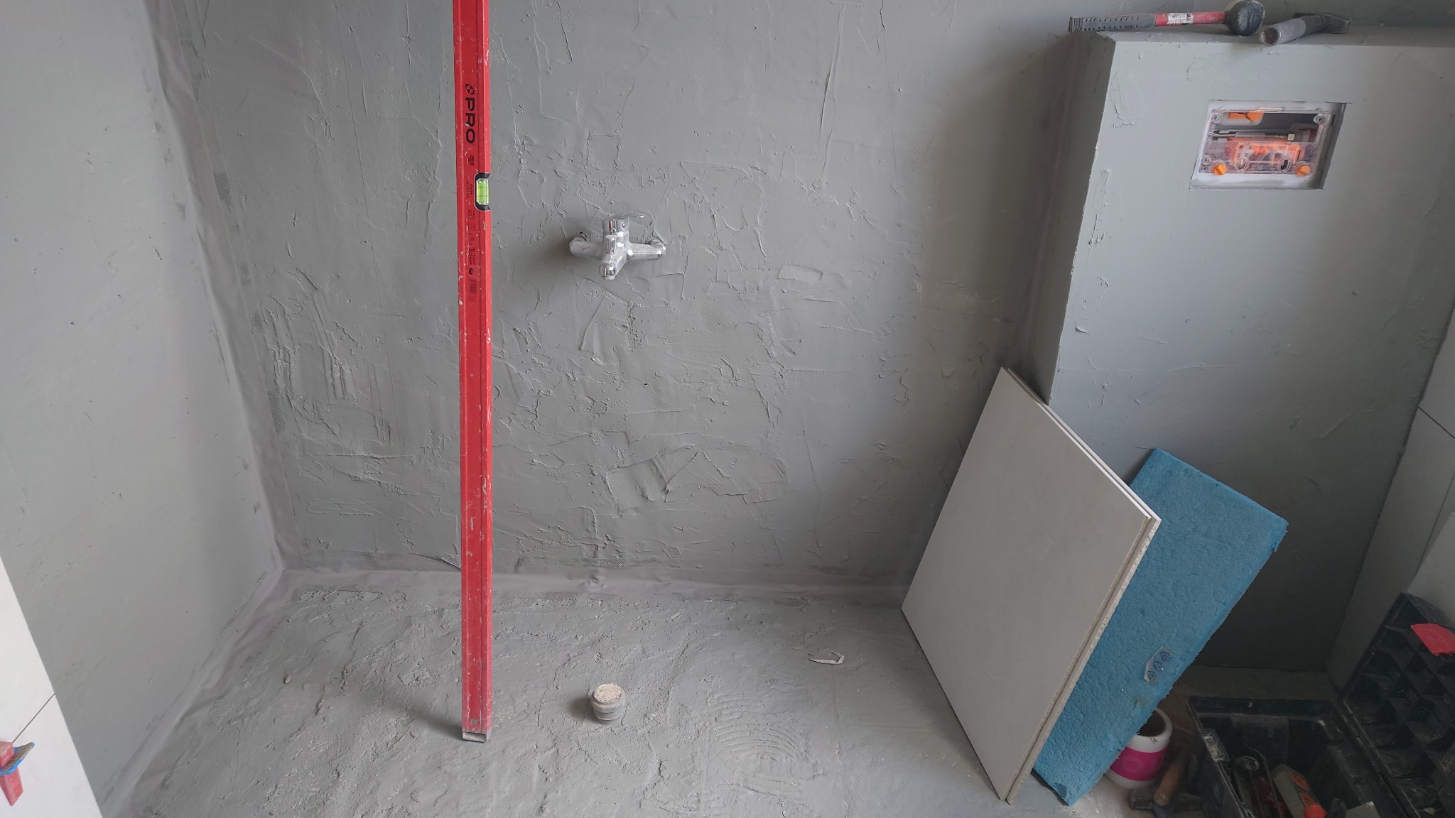 Surowa łazienka w trakcie prac: szare ściany z poziomnicą, widoczna instalacja wodna, płyty i narzędzia glazurnicze. Przygotowanie do dalszych prac wykończeniowych.