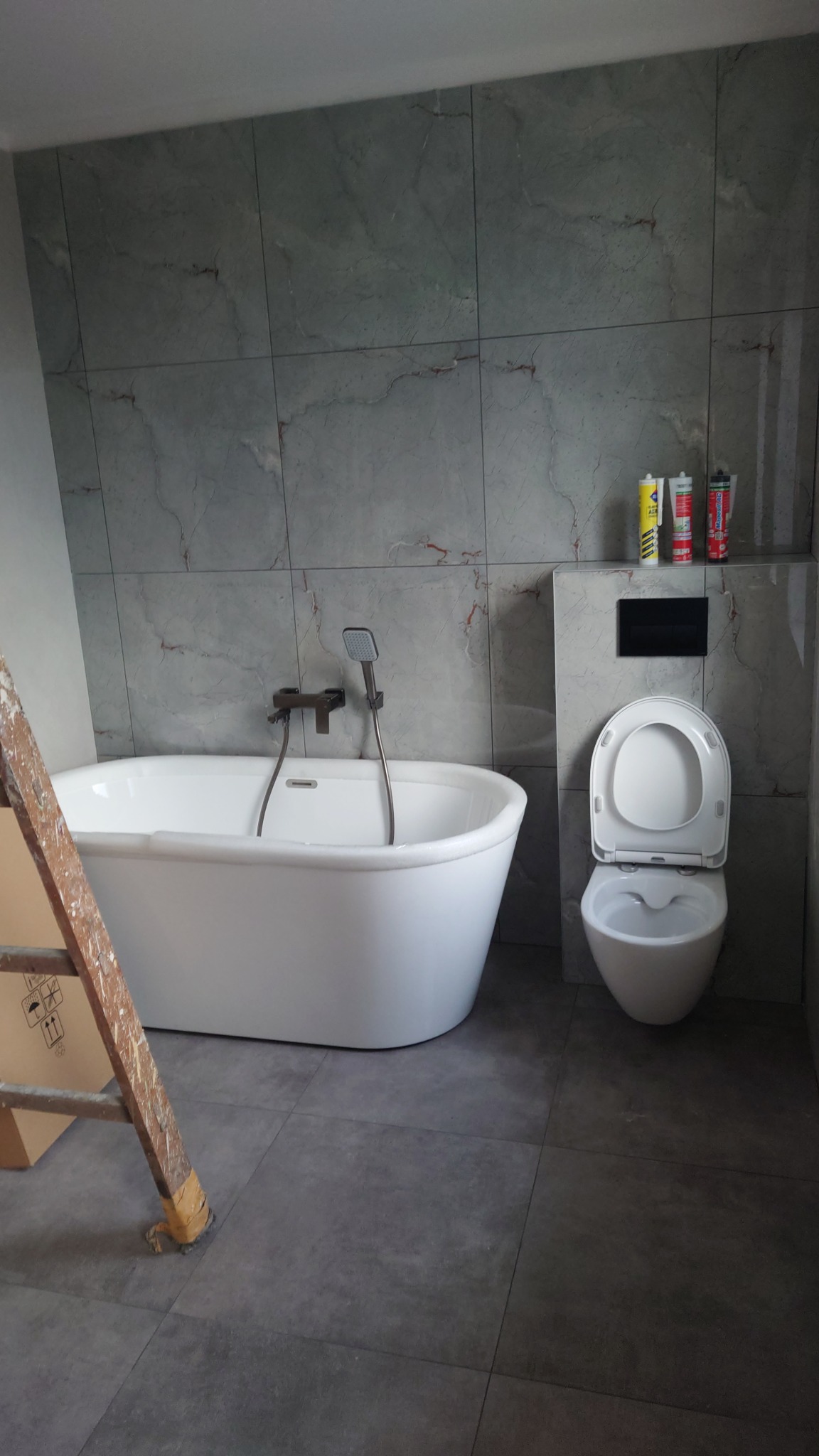 Nowoczesna łazienka w szarościach: wolnostojąca wanna, podwieszana toaleta, ściany i podłoga wyłożone płytkami imitującymi marmur. Widoczna drabina malarska.