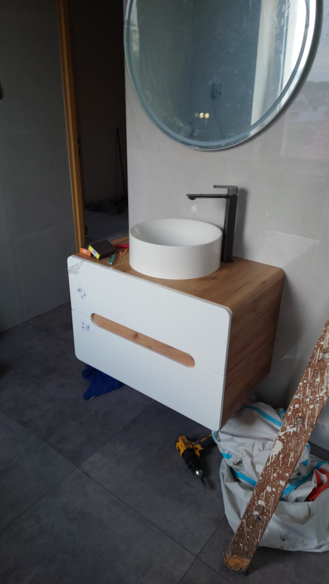 Nowoczesna umywalka nablatowa na szafce z drewnianym blatem i białymi frontami w łazience z szarą podłogą. Widoczne narzędzia i okrągłe lustro z podświetleniem.