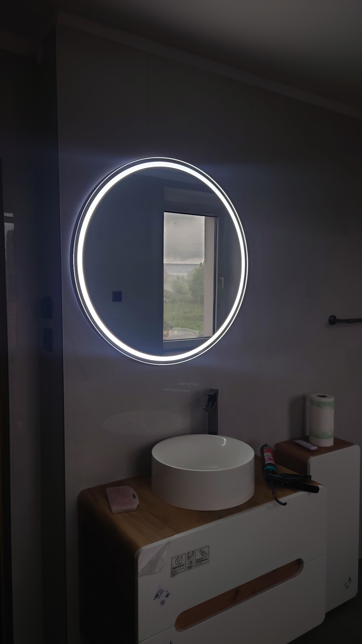 Nowoczesna łazienka: okrągłe lustro LED nad białą umywalką nablatową na szafce z drewnianym blatem. W tle widok z okna na zieleń.