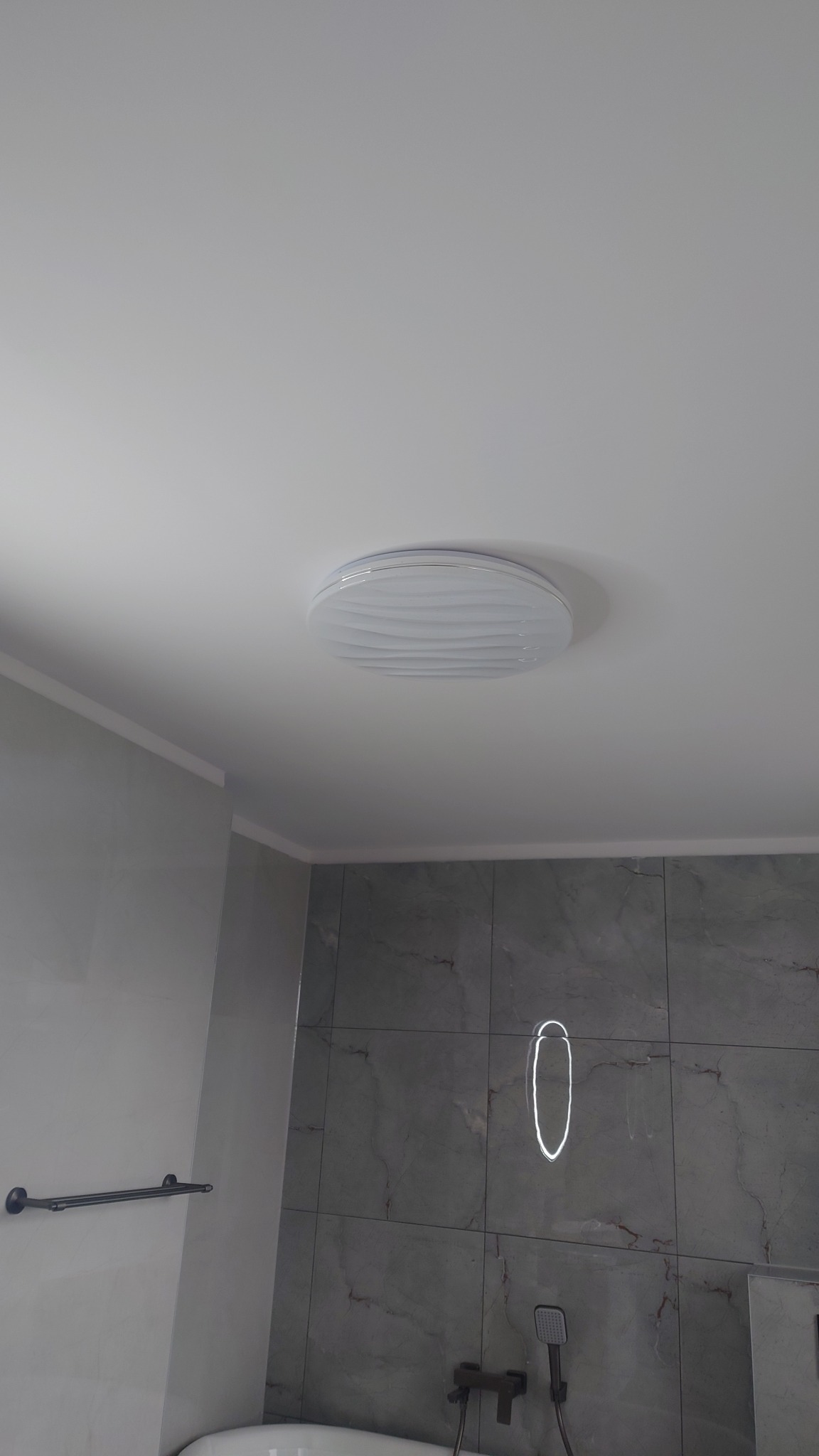 Wykończona łazienka z szarymi płytkami imitującymi marmur, okrągłą lampą sufitową i ciemną armaturą. Widoczny fragment wanny i wieszak na ręczniki.