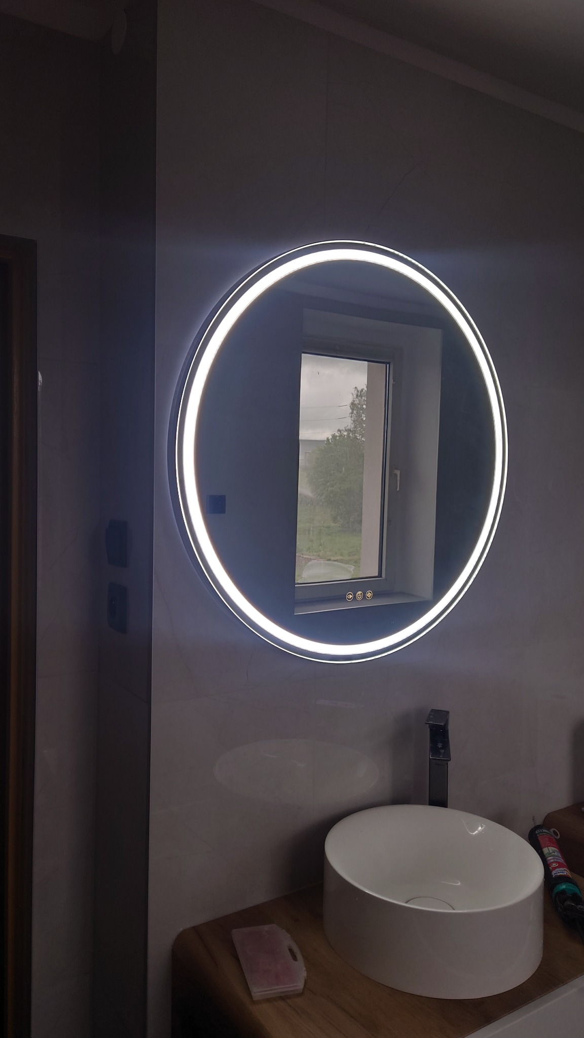 Okrągłe lustro LED nad umywalką w łazience, odbijające widok okna. Minimalistyczny design, jasne oświetlenie, drewniany blat, szare płytki na ścianie.