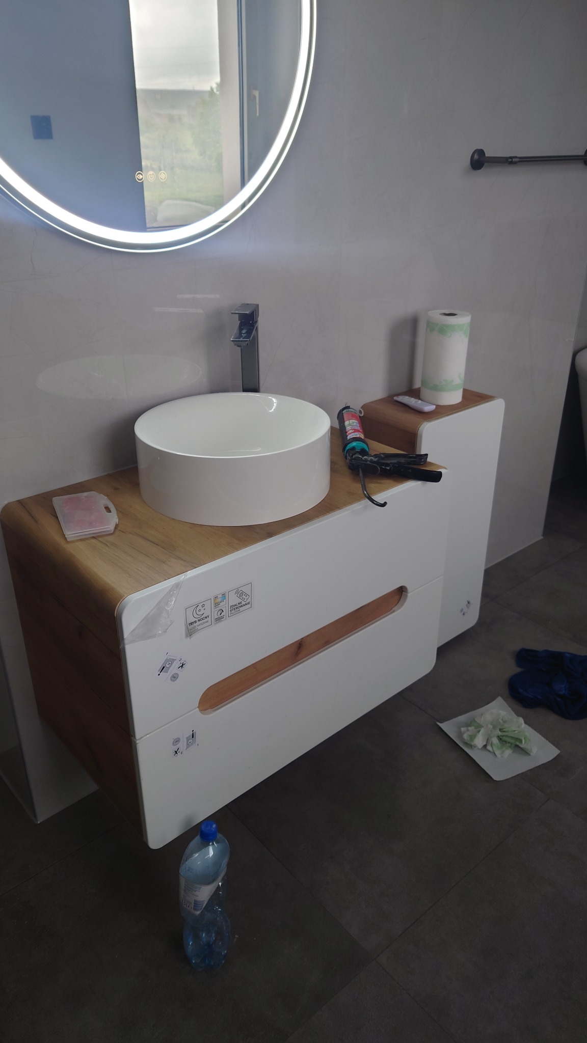 Nowoczesna umywalka nablatowa na szafce z drewnianym blatem i białymi frontami. Widoczny okrągły, podświetlany LED-ami lustro i narzędzia glazurnicze.
