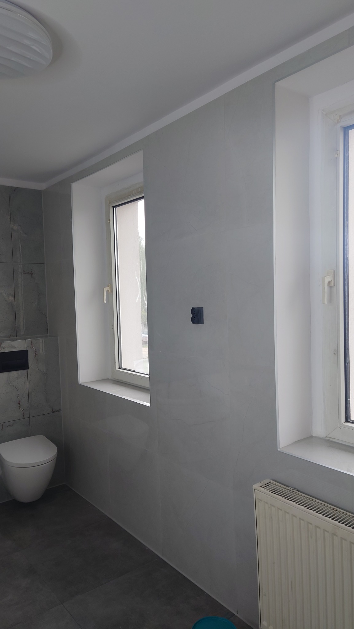 Wykończona łazienka w szarościach: płytki imitujące marmur na ścianach, grafitowa podłoga, biała toaleta podwieszana i okno z białymi ramami.