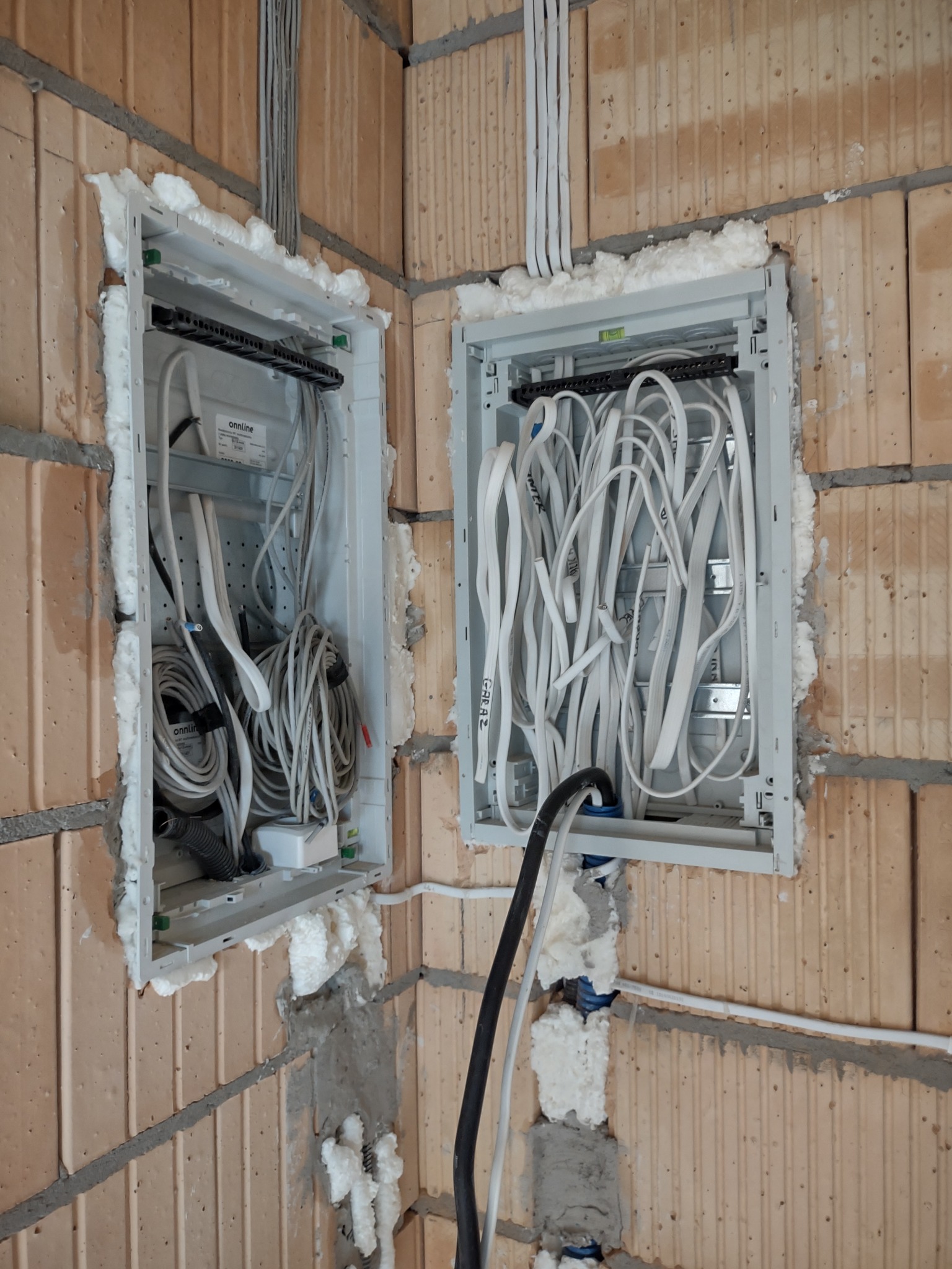 Dwie podtynkowe skrzynki elektryczne z kablami w trakcie montażu w ścianie z cegły, zabezpieczone pianką montażową.