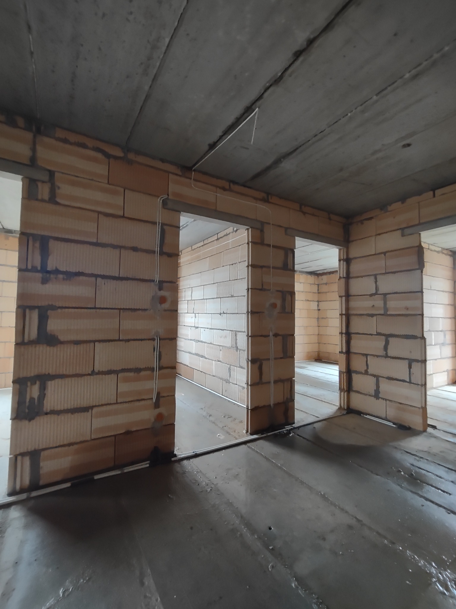Surowy stan wnętrza: ściany z cegły kratówki z widocznymi przewodami elektrycznymi, surowy betonowy sufit, mokra, wylana posadzka, widoczne otwory drzwiowe.