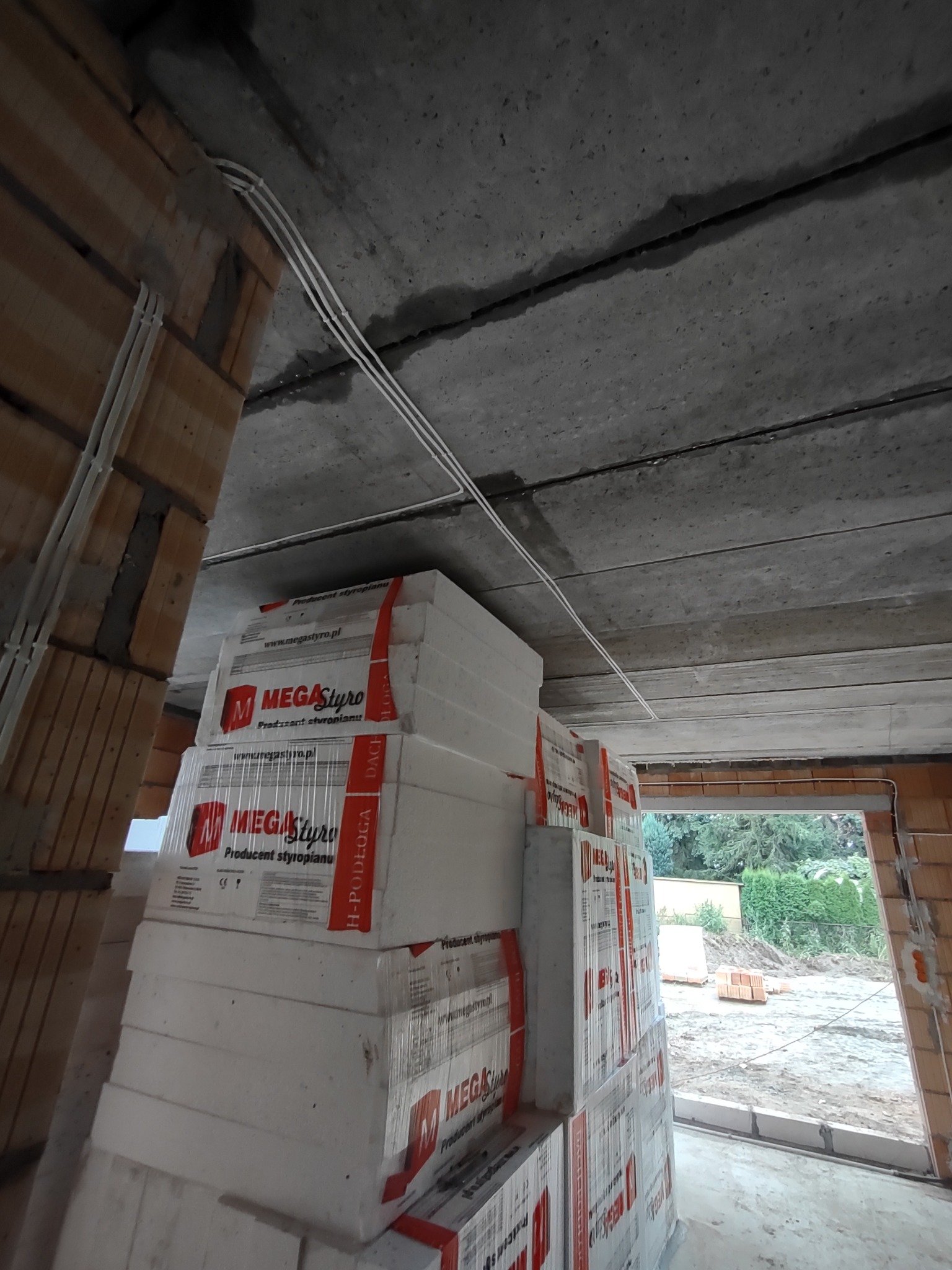 Wnętrze w trakcie budowy: ułożone paczki styropianu Mega Styro na podłogę, widoczne przewody elektryczne pod betonowym stropem i fragment ściany z pustaków ceramicznych, widok na zewnątrz przez...