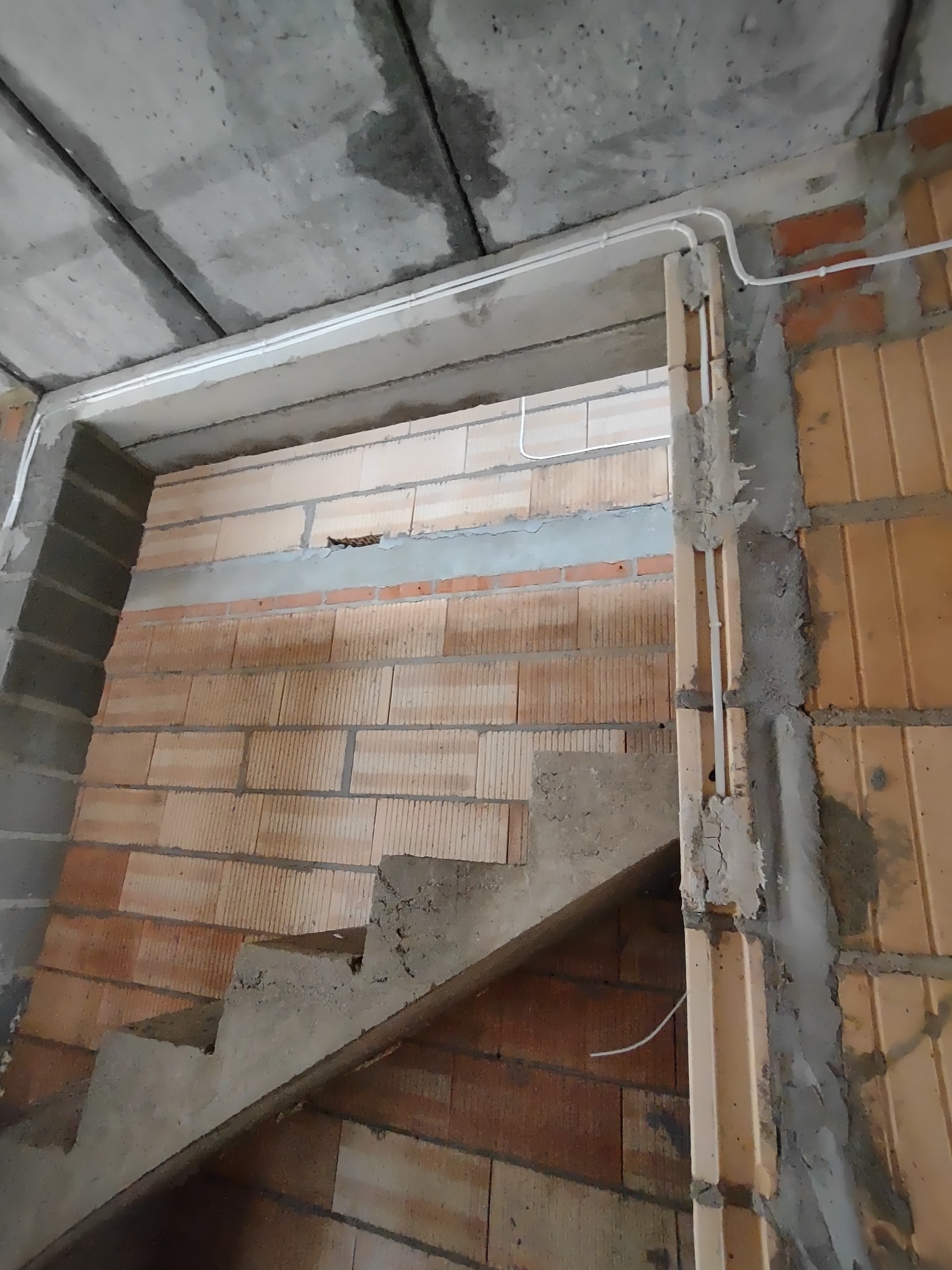 Surowy stan klatki schodowej w bloku z cegły, z widoczną instalacją elektryczną w białych rurkach PCV, sufit betonowy w tle.
