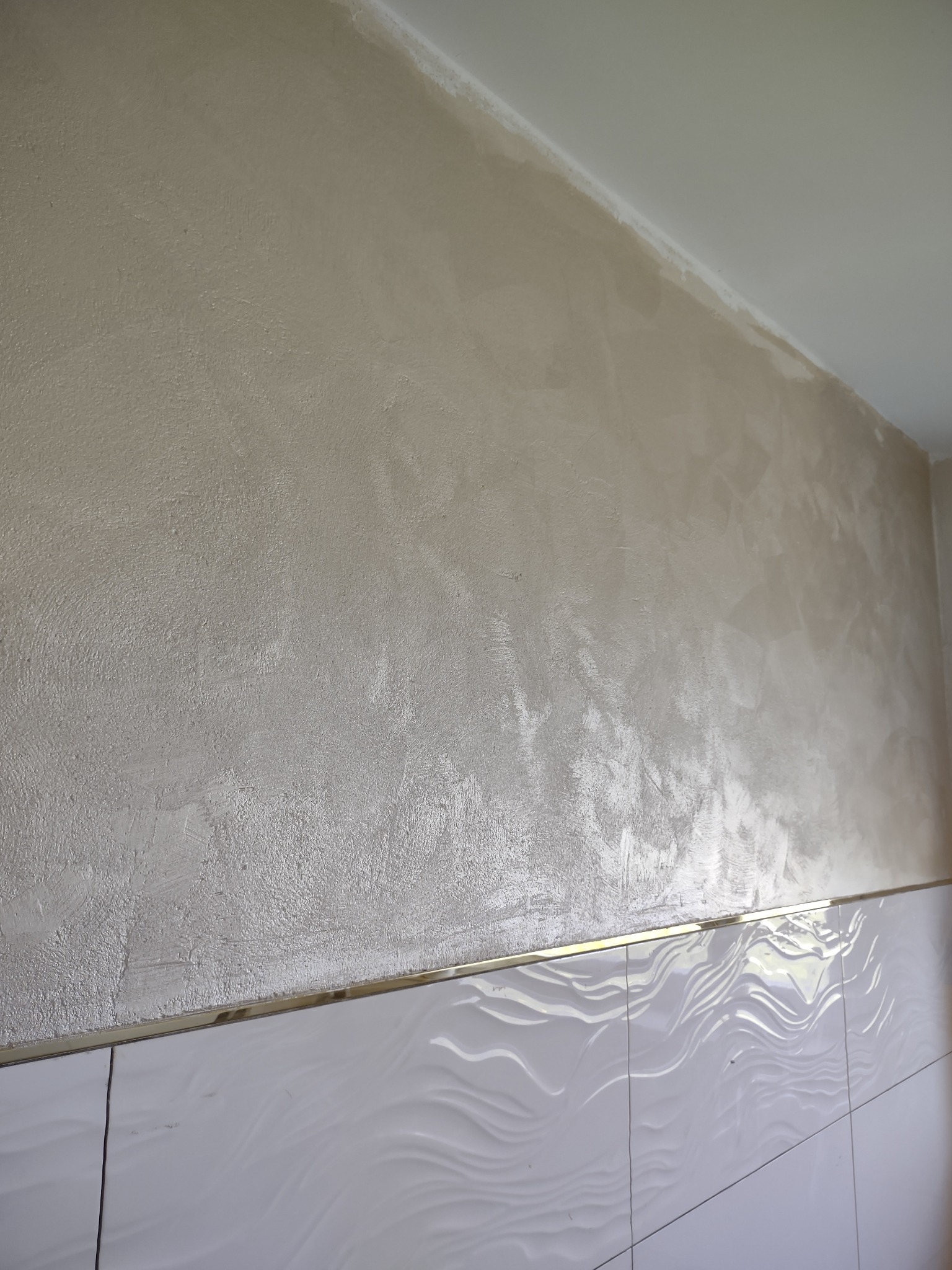 Ściana po szpachlowaniu z widoczną fakturą, poniżej białe płytki z falistym wzorem oddzielone złotą listwą.