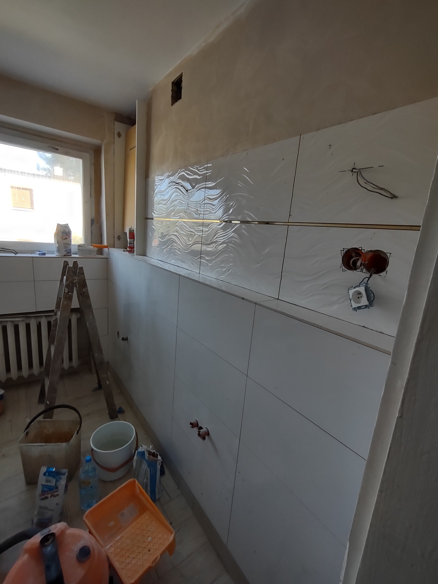 Wnętrze w trakcie remontu: częściowo ułożone białe płytki z falistym wzorem na ścianie, widoczne puszki instalacyjne, drabina oparta o ścianę, wiadra i narzędzia na podłodze.