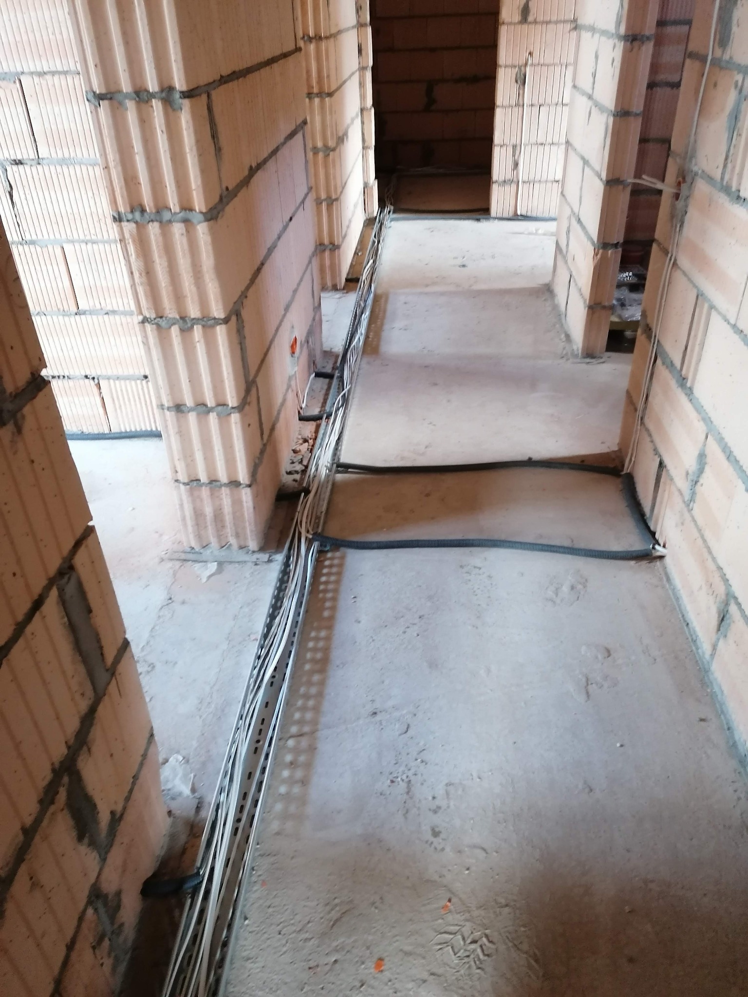 Wnętrze w trakcie budowy z widocznymi przewodami elektrycznymi ułożonymi wzdłuż ściany z cegły ceramicznej dziurawki, betonowa podłoga w surowym stanie.