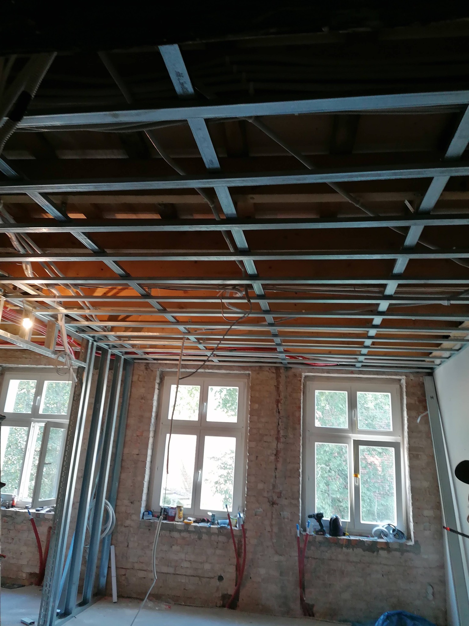 Metalowy stelaż sufitu podwieszanego w trakcie montażu, widoczne przewody elektryczne i fragmenty instalacji, surowa cegła na ścianach z trzema oknami.
