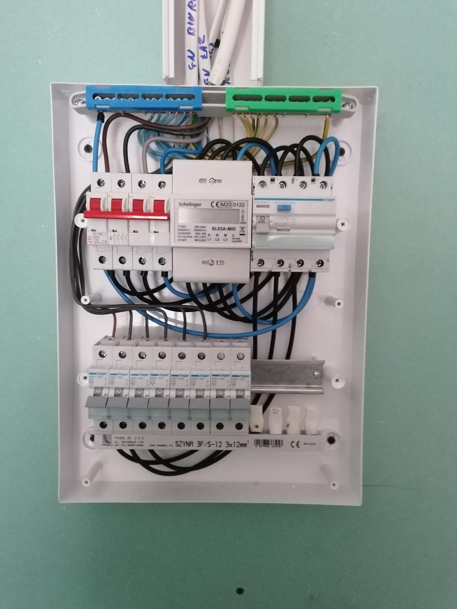 Rozdzielnica elektryczna z zainstalowanymi bezpiecznikami, przewodami i szynami, zamontowana na ścianie pomalowanej na zielono.