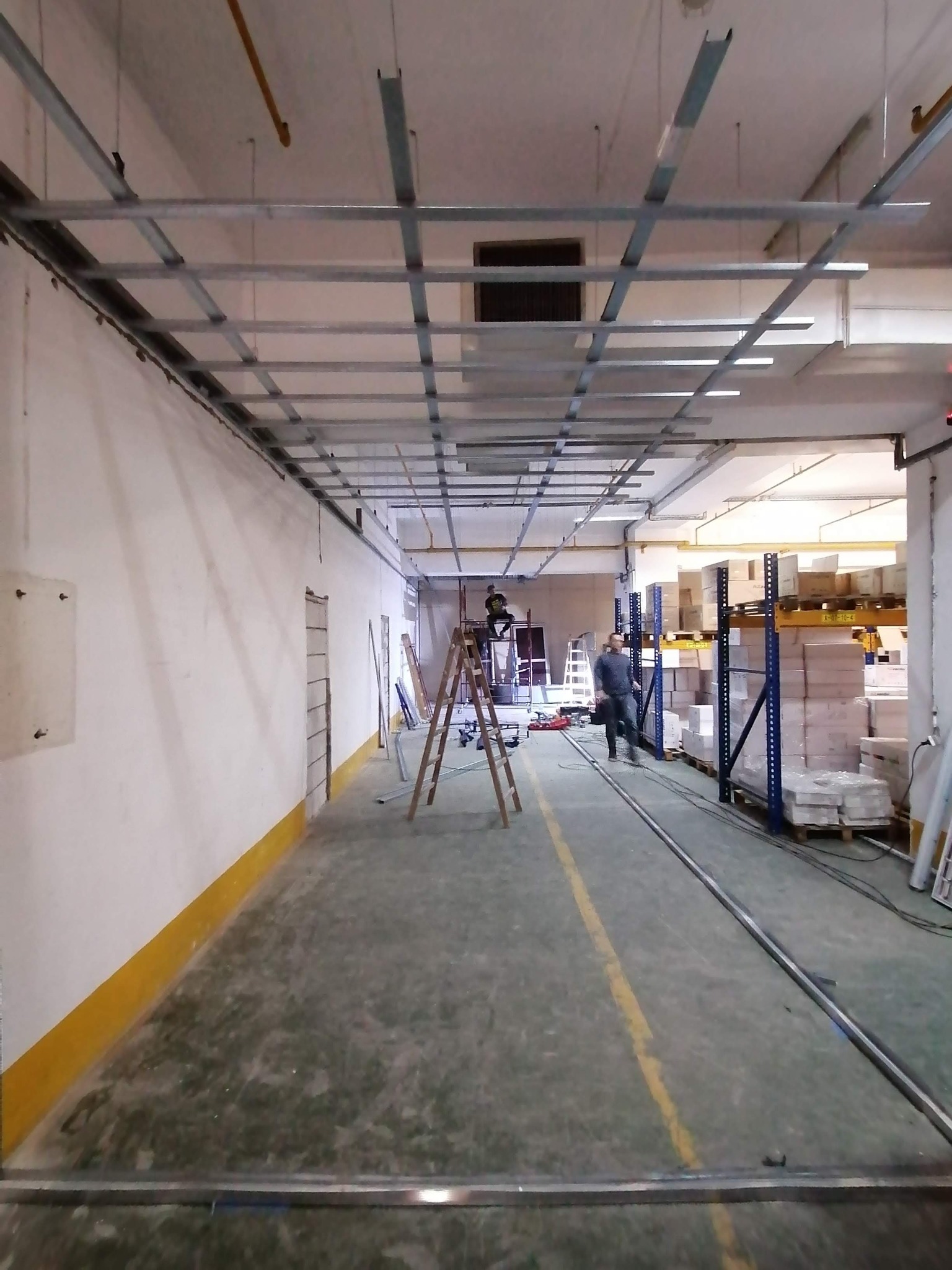 Montaż metalowej konstrukcji sufitu podwieszanego w magazynie, widoczni dwaj pracownicy na drabinach, w tle regały z towarem.