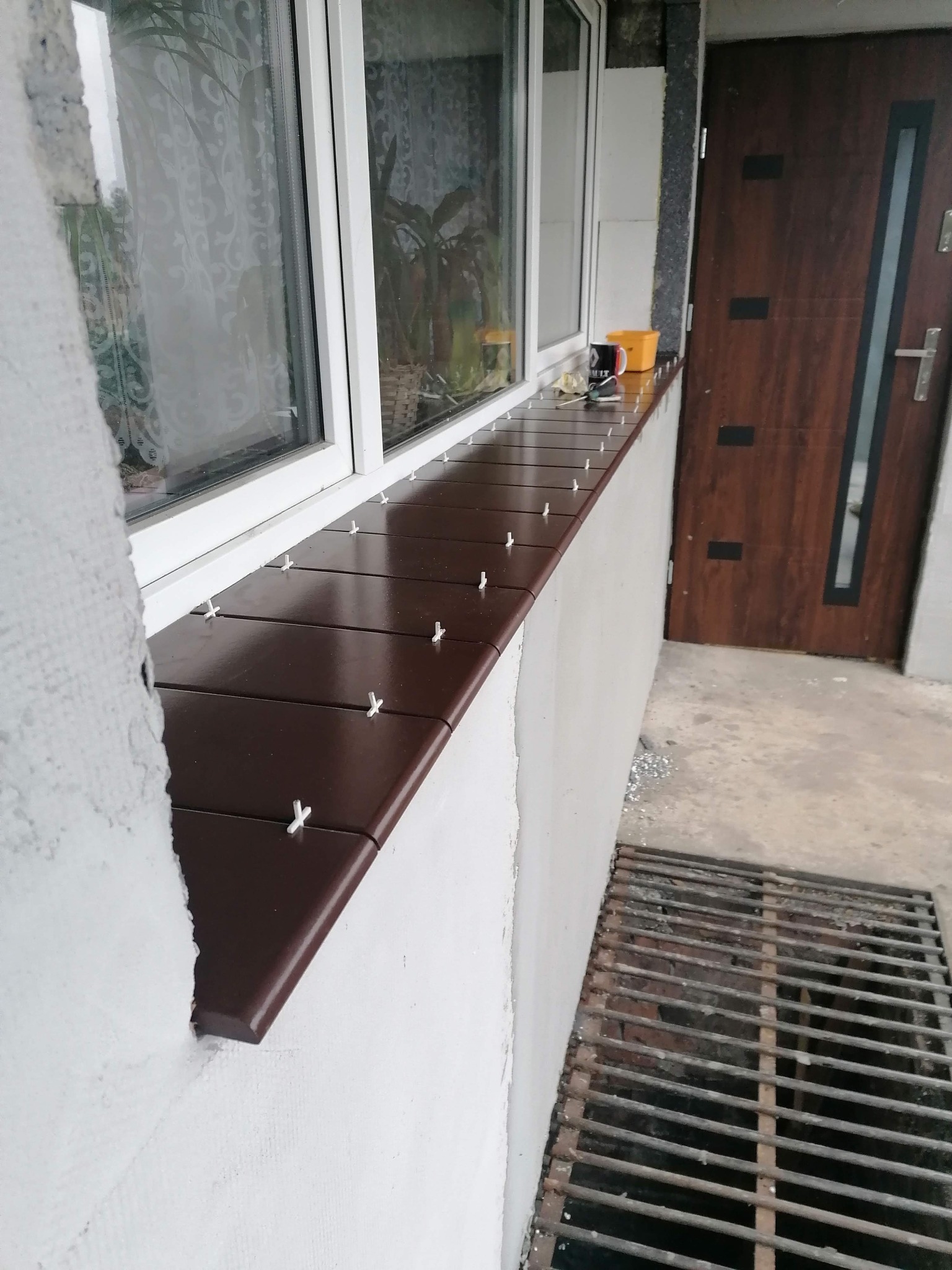 Montaż brązowych płytek parapetowych zewnętrznych z białymi krzyżykami dystansowymi, widoczna okleina okienna i fragment elewacji z niedokończonym tynkiem.