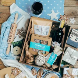 Świąteczny kosz prezentowy z winem, kawą, pierniczkami, drewnianym wałkiem z motywem reniferów, kalendarzem adwentowym i dekoracjami w kształcie gwiazd na drewnianym stole przykrytym niebieską...