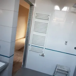 Wnętrze łazienki w trakcie remontu: białe płytki z niebieskim paskiem, grzejnik drabinkowy, widoczne kable elektryczne wystające ze ściany, oraz fragment sąsiedniego pomieszczenia z drewnianą podłogą.