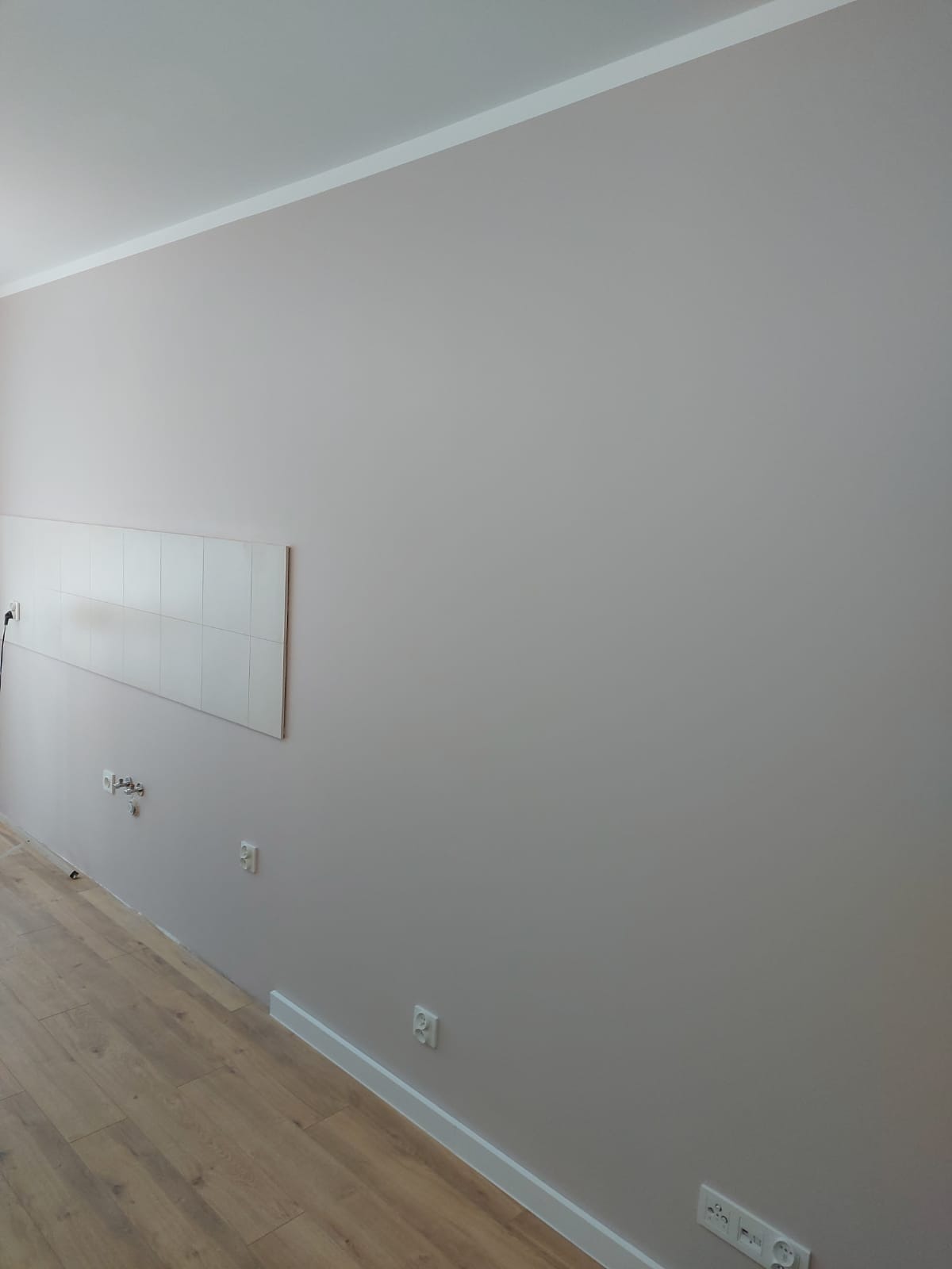 Świeżo pomalowana ściana w jasnoszarym odcieniu z widocznymi gniazdkami elektrycznymi i fragmentem drewnianej podłogi, listwy przypodłogowe w kolorze białym, na ścianie położone białe płytki...