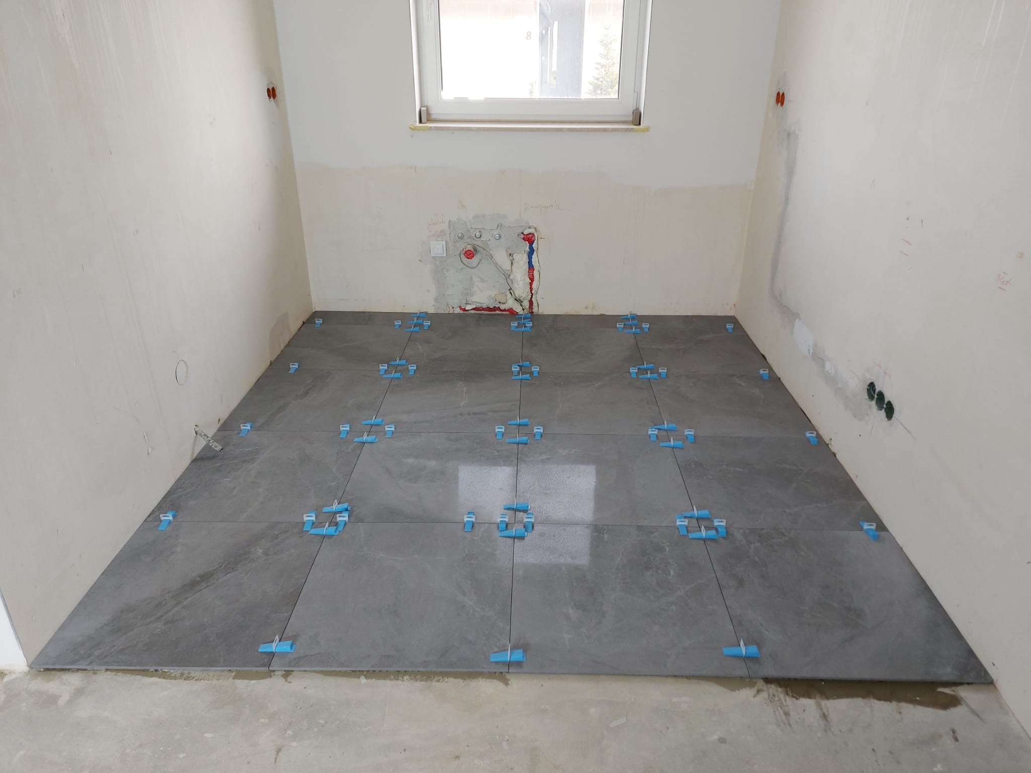 Układanie szarych płytek podłogowych w łazience, widoczne nieotynkowane ściany i niebieskie krzyżyki dystansowe.