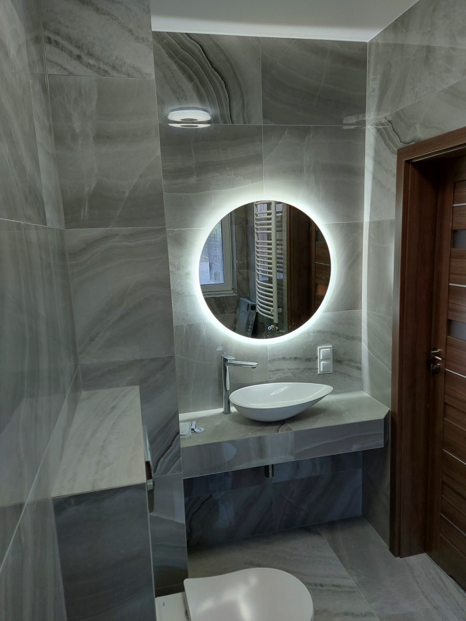 Nowoczesna łazienka z szarymi płytkami imitującymi marmur, okrągłym lustrem z podświetleniem LED, owalną białą umywalką i drewnianymi drzwiami.