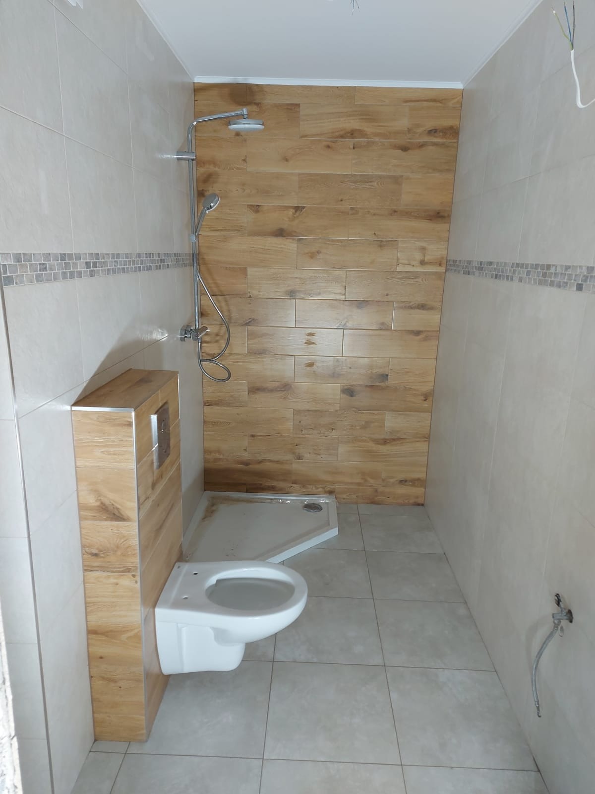 Wykończona łazienka z kabiną prysznicową, podwieszaną toaletą i ścianą wyłożoną płytkami imitującymi drewno, z dekoracyjnym pasem płytek mozaikowych pod sufitem.