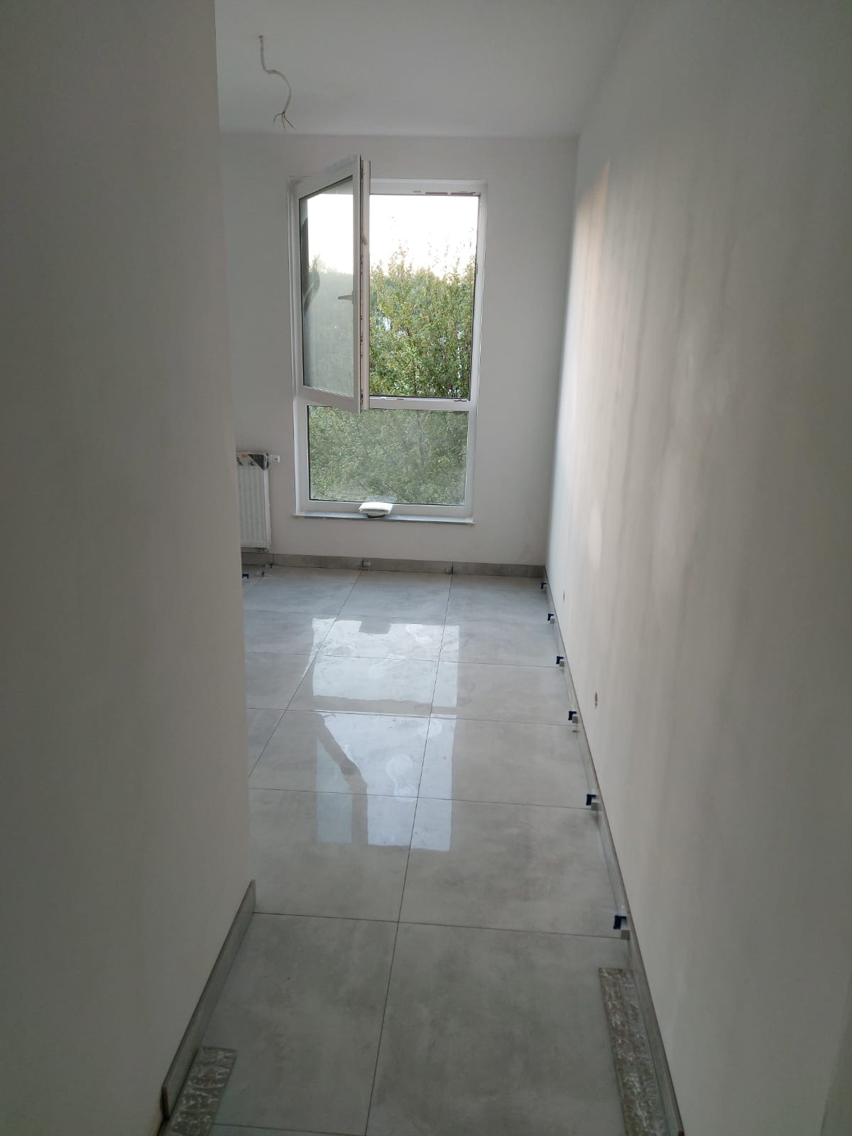 Układanie jasnoszarych płytek podłogowych w nowym pomieszczeniu, widoczne krzyżyki dystansowe, otwarte okno z widokiem na zieleń.