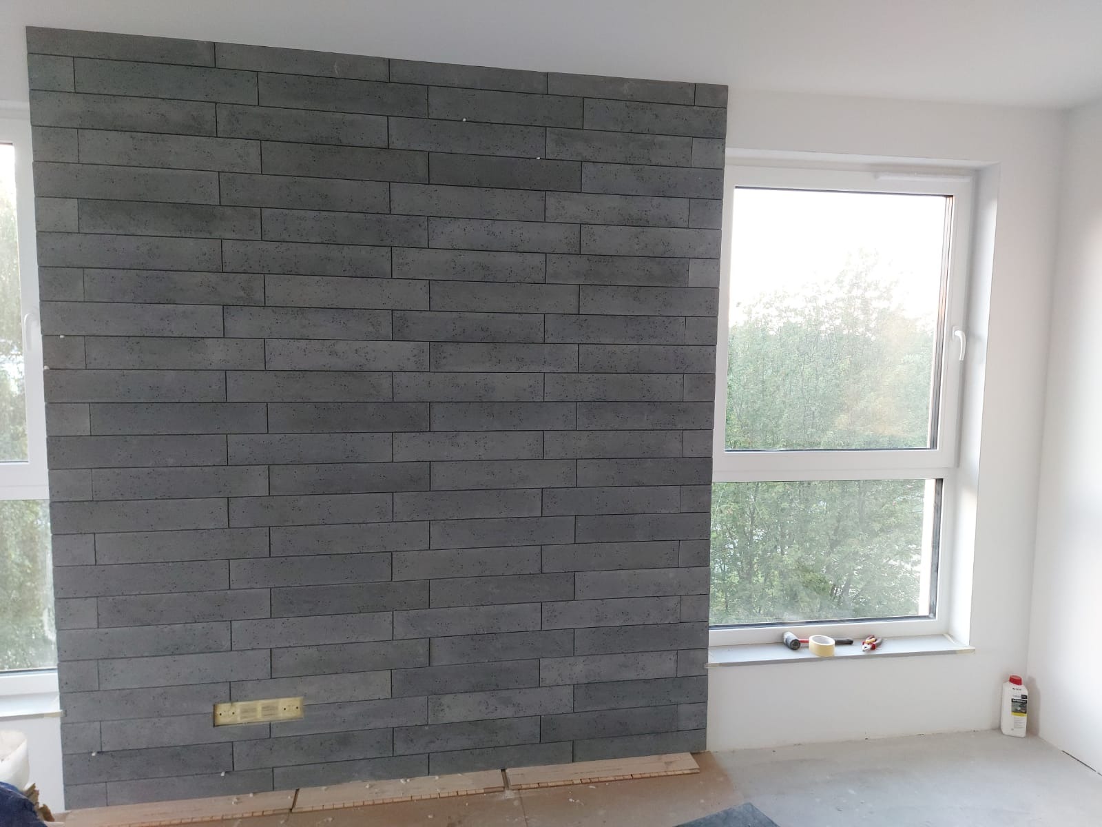 Ściana wyłożona szarą cegłą dekoracyjną w trakcie montażu, widoczne puszki elektryczne, okno z widokiem na drzewa i narzędzia na parapecie.