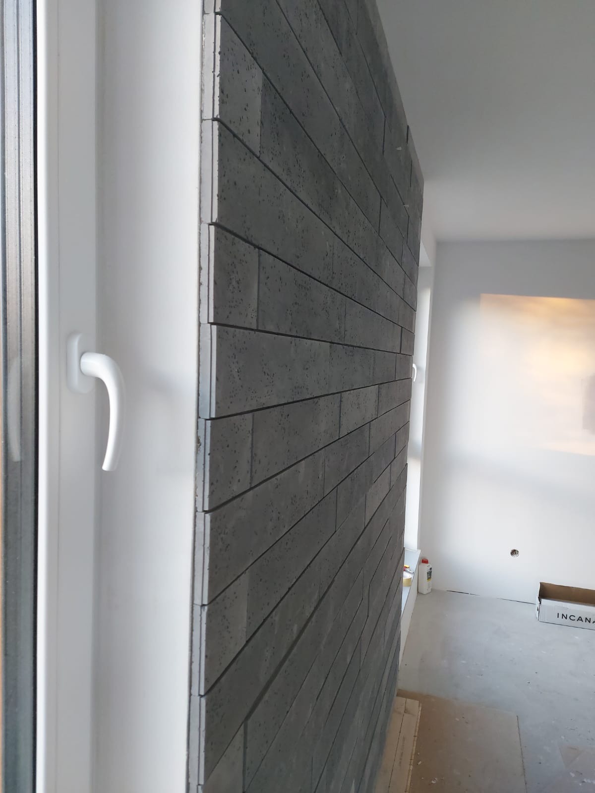 Fragment ściany wykończonej szarą cegłą dekoracyjną, widok z boku, obok okno PCV, w tle białe ściany w trakcie prac wykończeniowych.