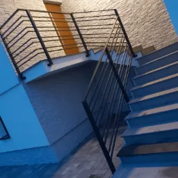 Metalowa balustrada schodowa z elementami spawanymi w Jaktorowie, widok z dołu na schody zewnętrzne i elewację budynku.