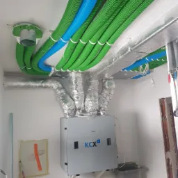 Centrala wentylacyjna z rekuperacją KCX zamontowana w pomieszczeniu, widoczne rury wentylacyjne w kolorze zielonym i niebieskim oraz izolowane przewody doprowadzające powietrze.