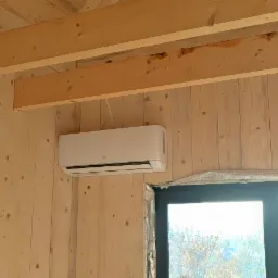 Biały klimatyzator ścienny marki AUX zamontowany w pomieszczeniu wykończonym jasnym drewnem, widok z okna na zewnątrz.