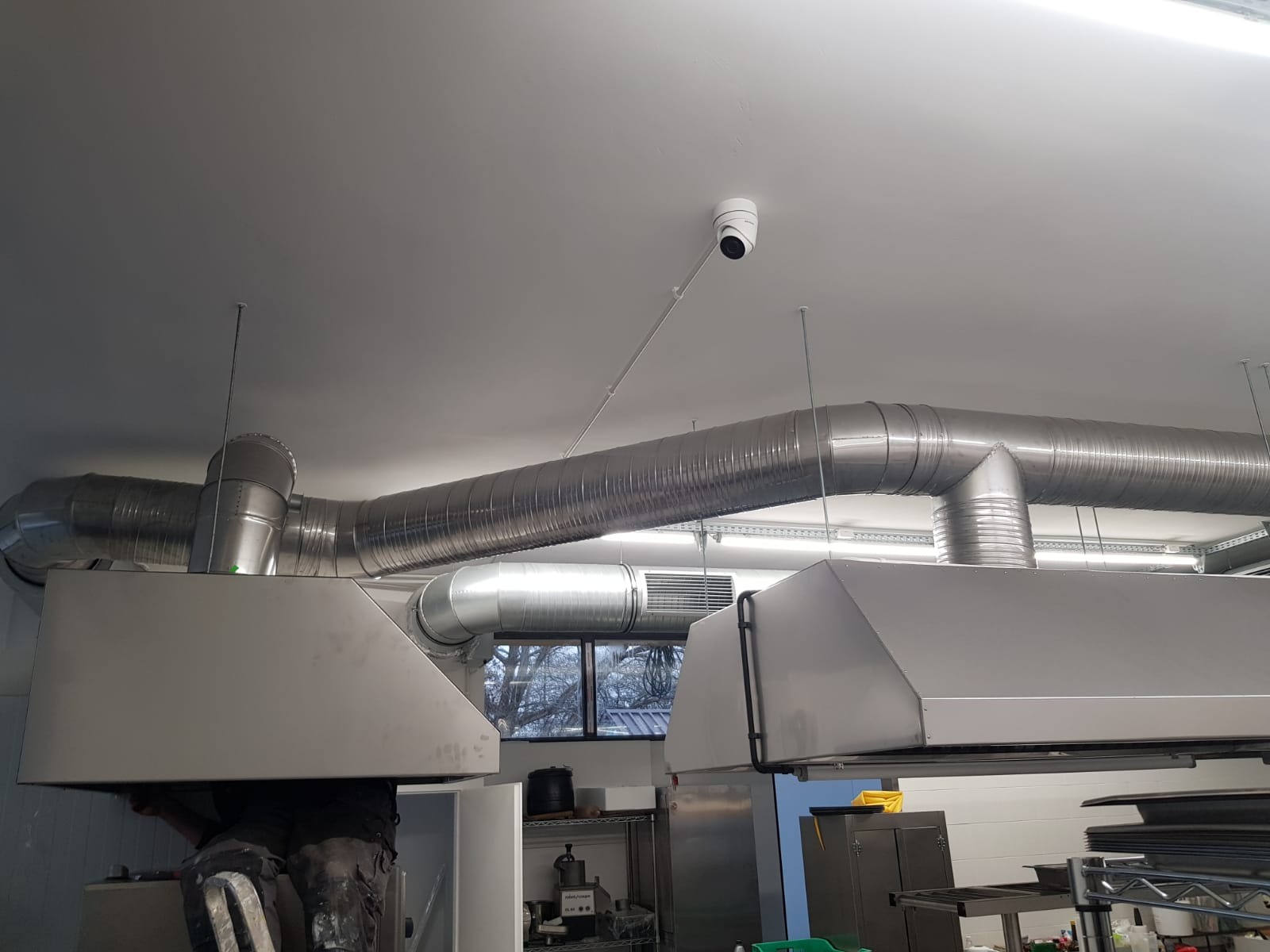 Montaż przemysłowej instalacji wentylacyjnej z okrągłych kanałów i okapów w kuchni, widoczna kamera monitoringu.