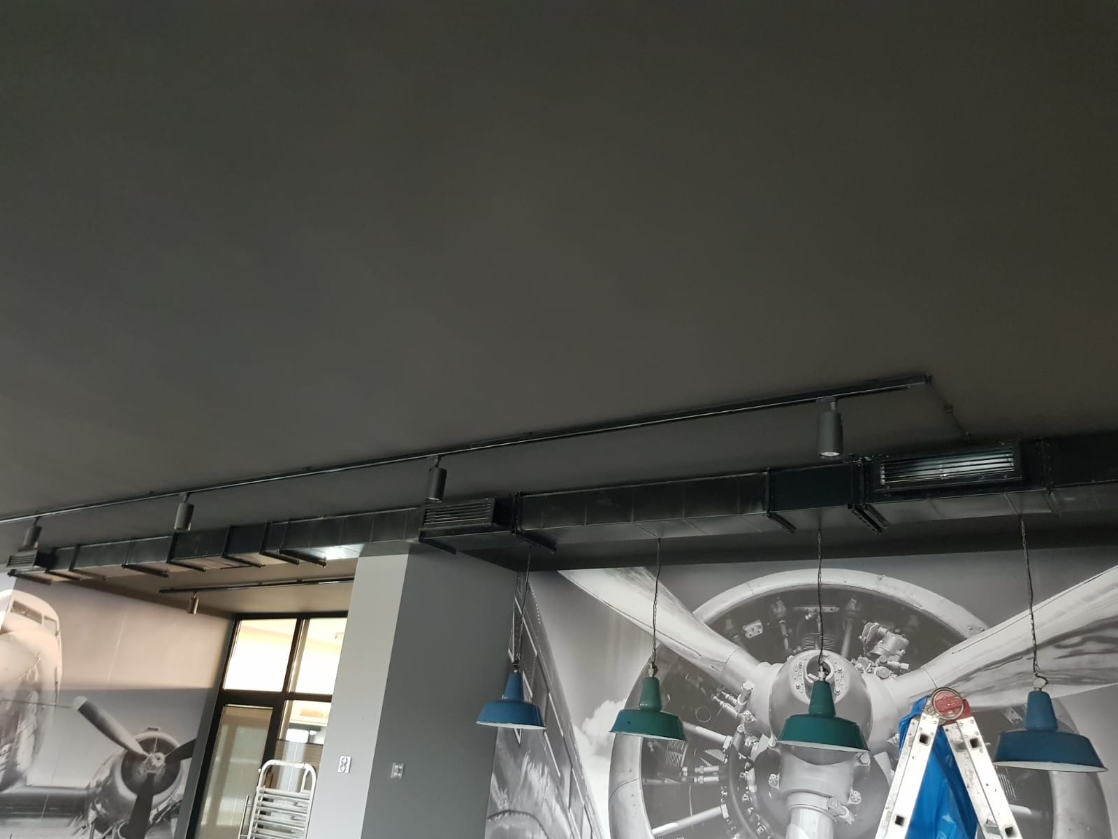 Czarny system kanałów wentylacyjnych pod sufitem w pomieszczeniu z dekoracją ściany w stylu lotniczym, widoczna drabina i wiszące lampy.